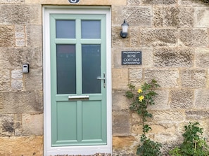 Eine Steinsäule mit einer grünen Tür, einem 'ROSE COTTAGE'-Schild und einer kleinen Pflanze mit gelben Blüten.