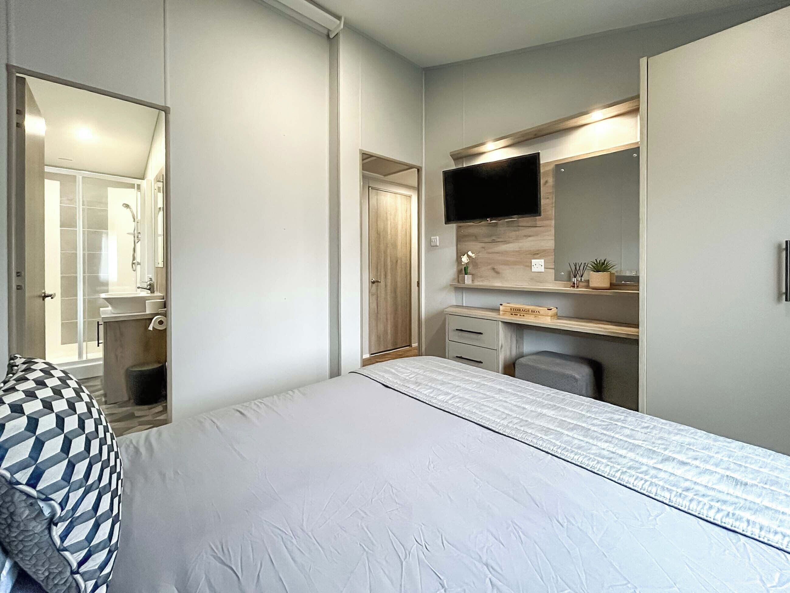 Une chambre à coucher avec un lit, un bureau, une chaise, une télévision et une salle de bain visible à travers une porte ouverte.