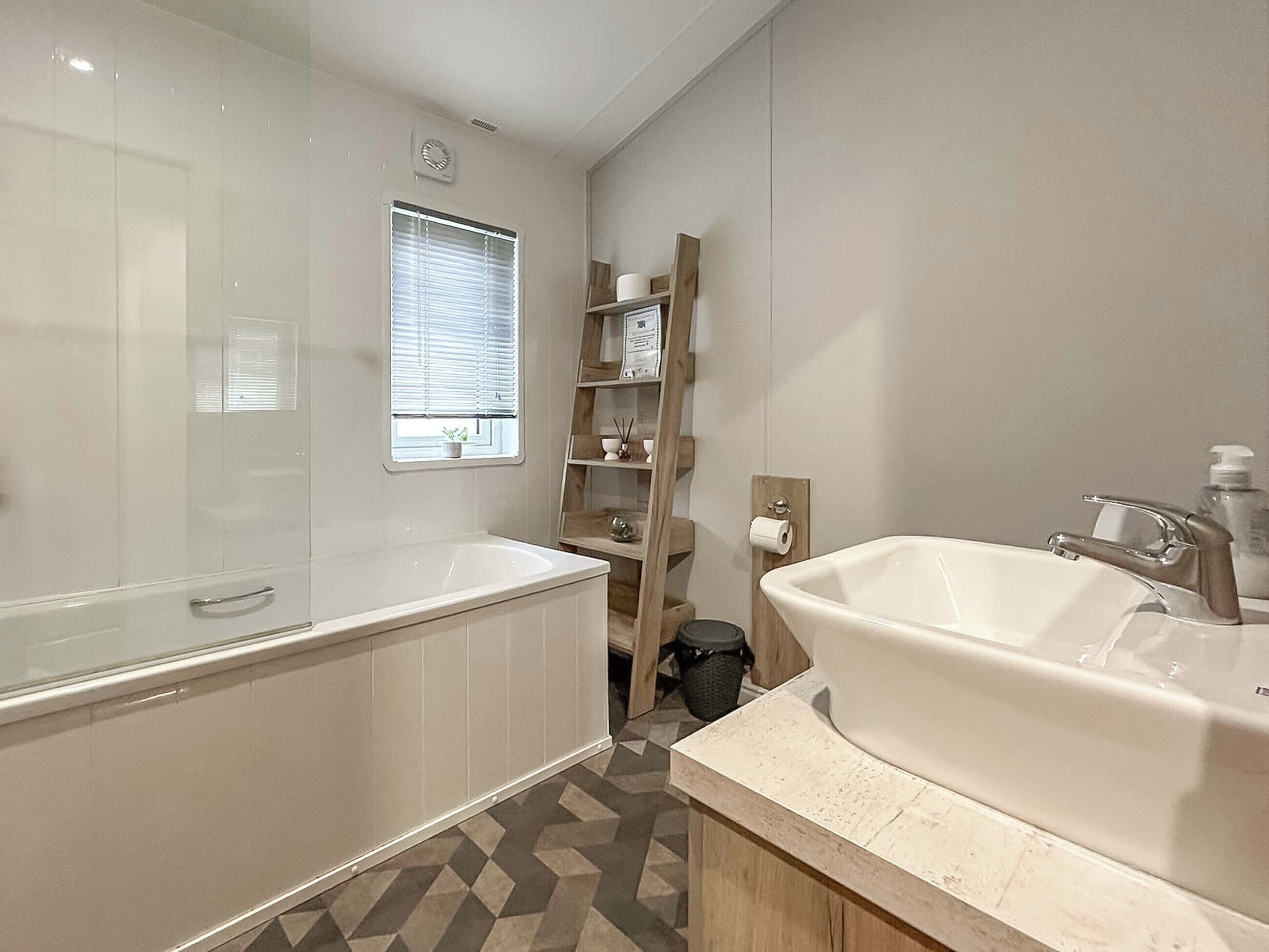 Une salle de bain moderne avec une grande baignoire, un lavabo séparé, une fenêtre avec des stores et une petite étagère contenant des articles de toilette.