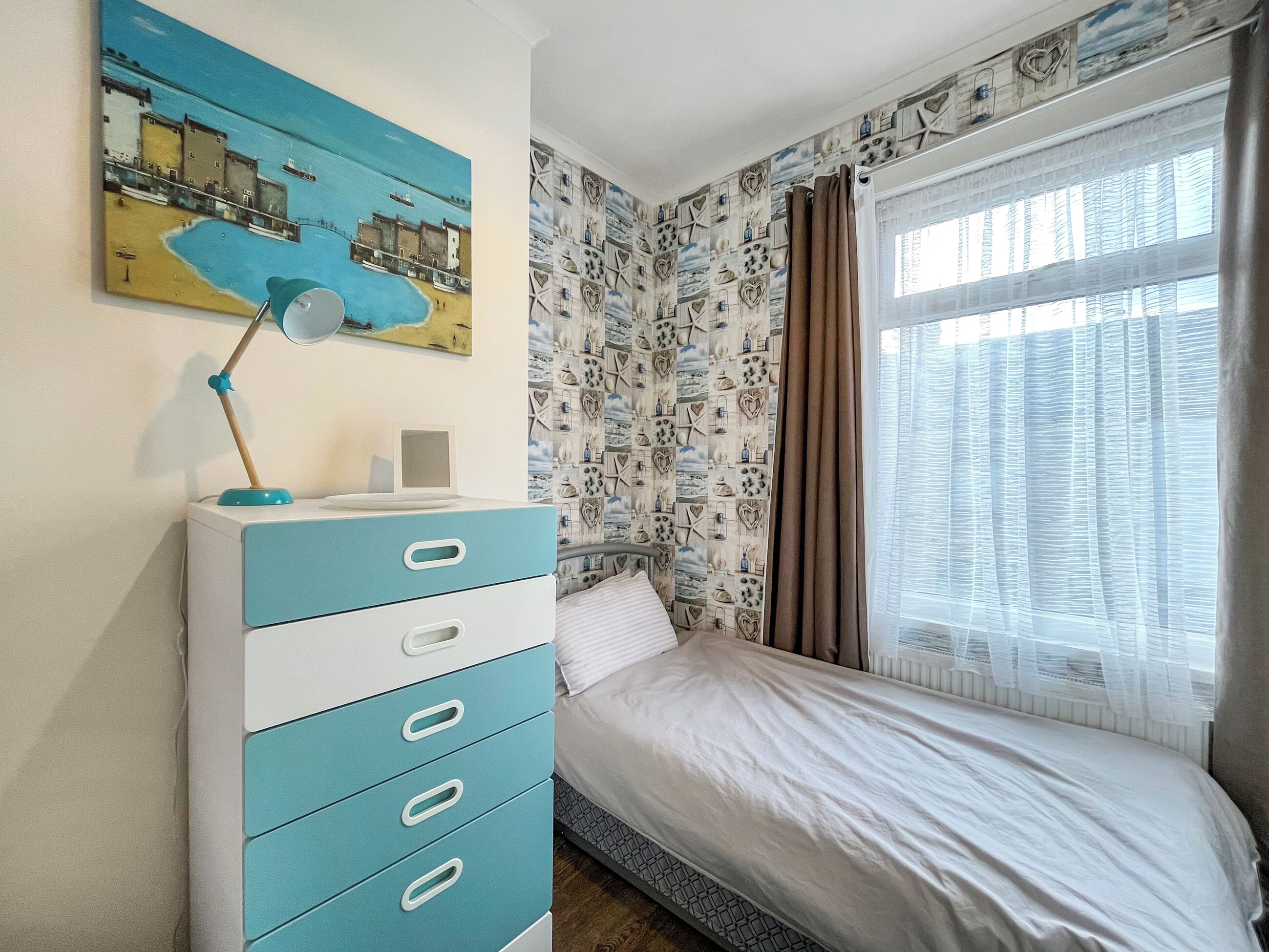 A bedroom with a bed, a dresser, a desk lamp, a painting, and a window with curtains.