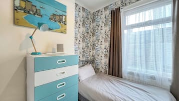 A bedroom with a bed, a dresser, a desk lamp, a painting, and a window with curtains.