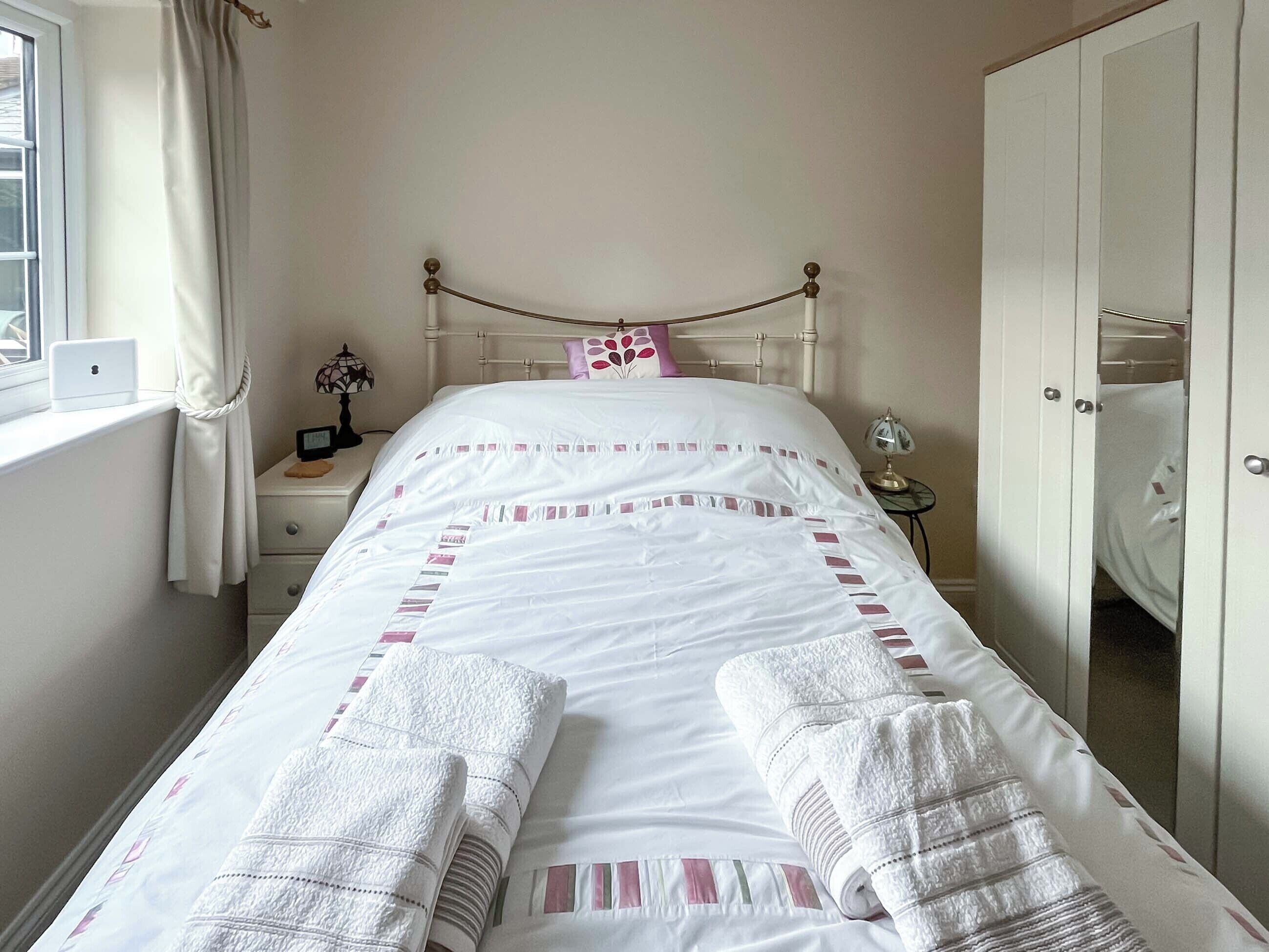 A neatly made bed with white bedding and pink accents, a bedside lamp, a laptop on a nightstand, and a wardrobe.
