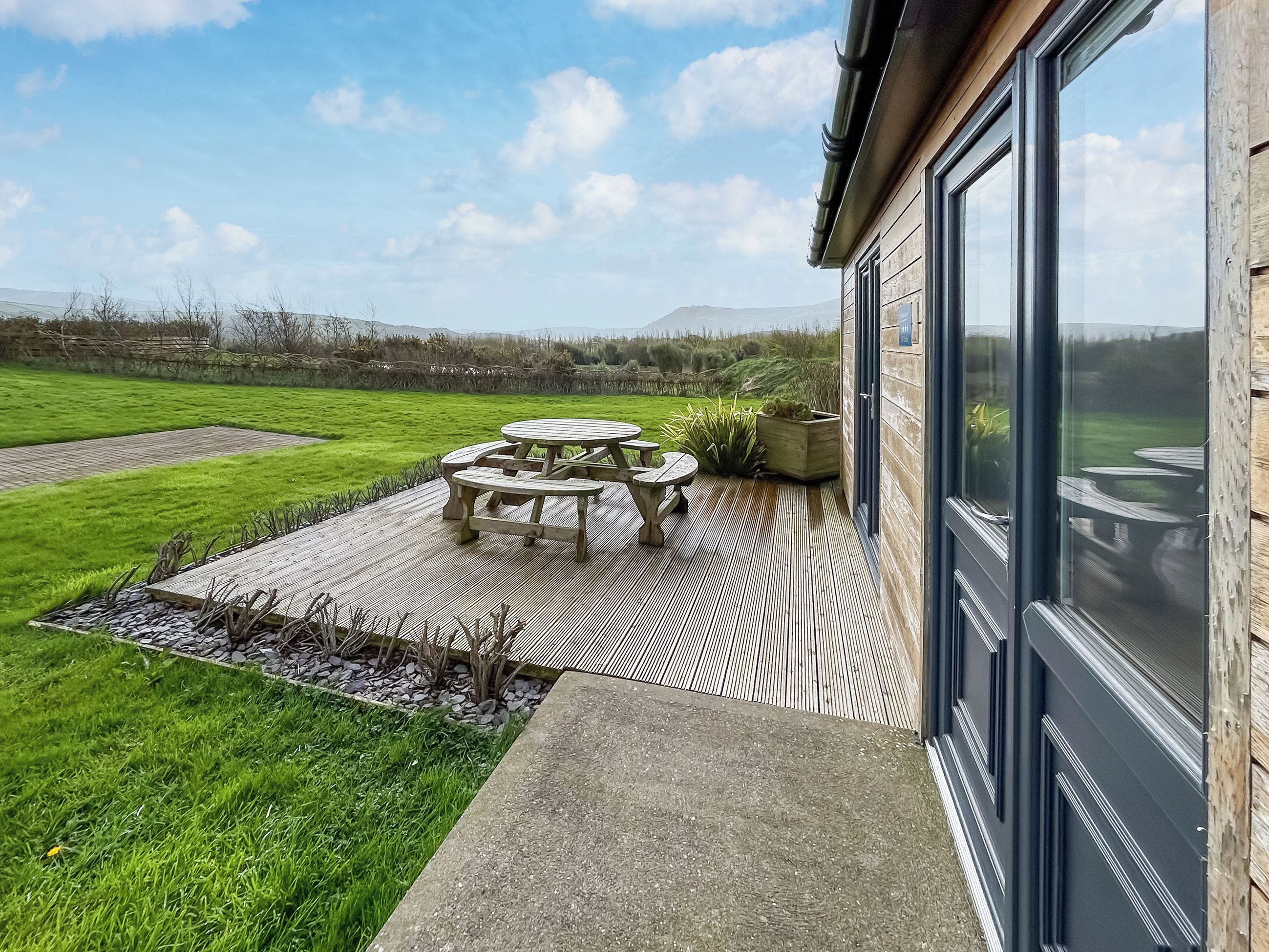 Eine Terrasse mit einem Holztisch und -stühlen, einem Kiesweg und Blick auf ein grünes Feld und ferne Hügel.