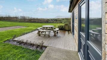 Eine Terrasse mit einem Holztisch und -stühlen, einem Kiesweg und Blick auf ein grünes Feld und ferne Hügel.