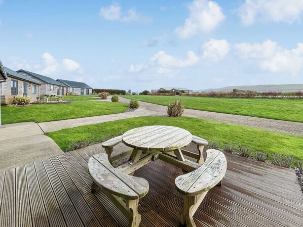 A wooden deck with a picnic table and benches, surrounded by a well-maintained lawn and a building in the background.