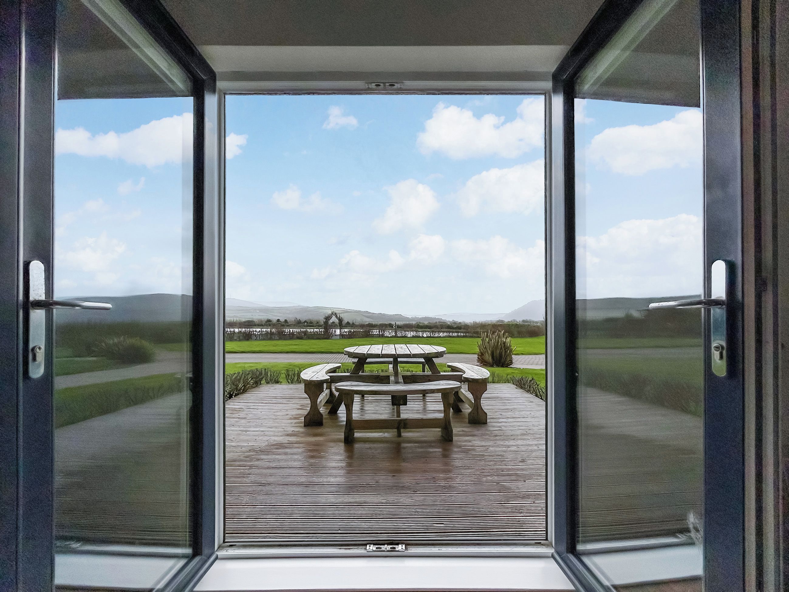 A view through a glass door of a patio area with a wooden table and chairs, a grassy field, and distant hills.