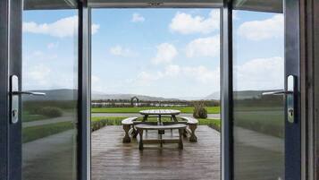 A view through a glass door of a patio area with a wooden table and chairs, a grassy field, and distant hills.