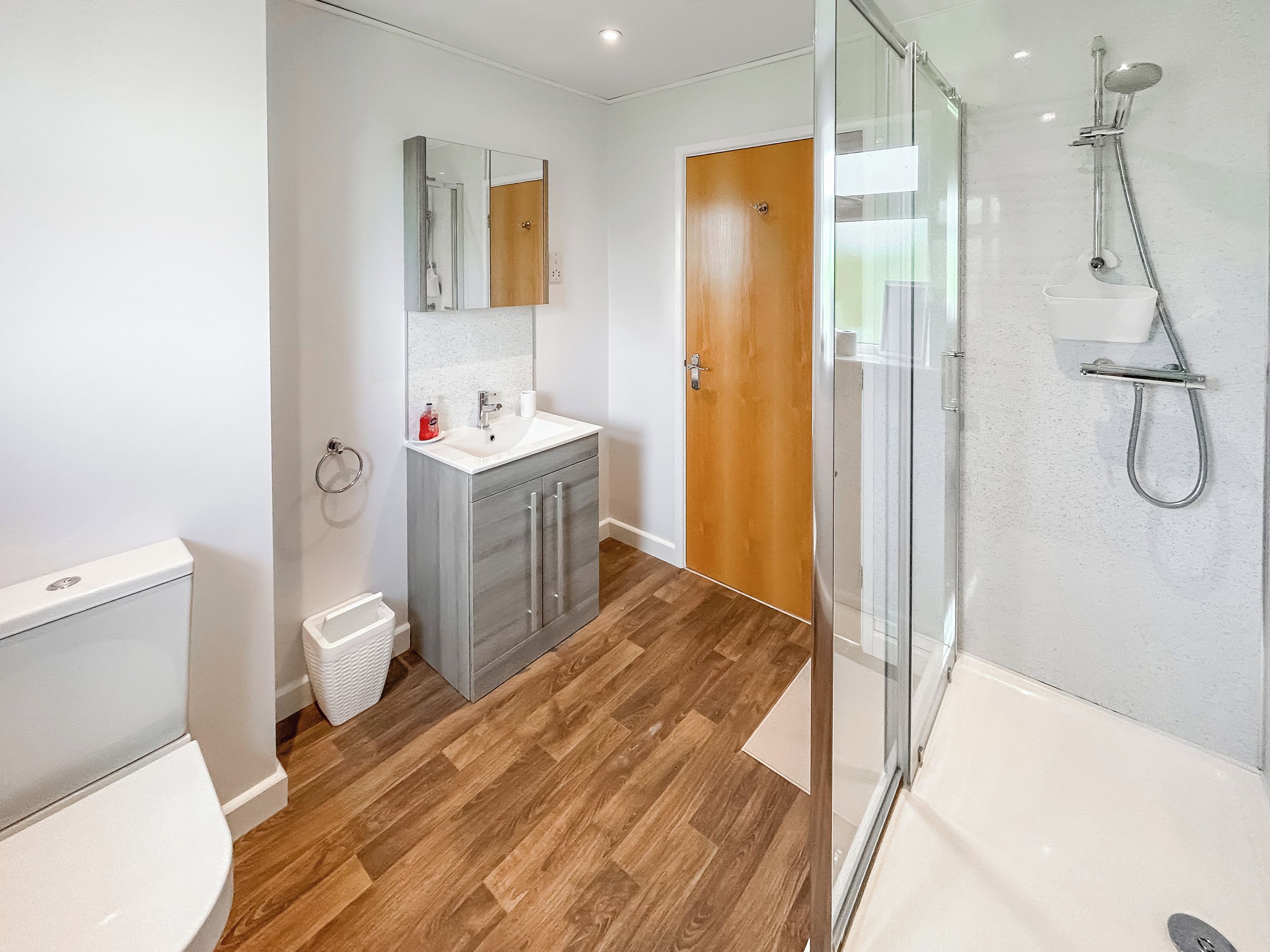A modern bathroom with a wooden floor, a glass shower enclosure, a white sink, and a wooden door.