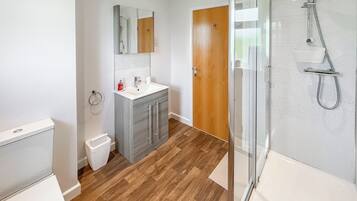 A modern bathroom with a wooden floor, a glass shower enclosure, a white sink, and a wooden door.