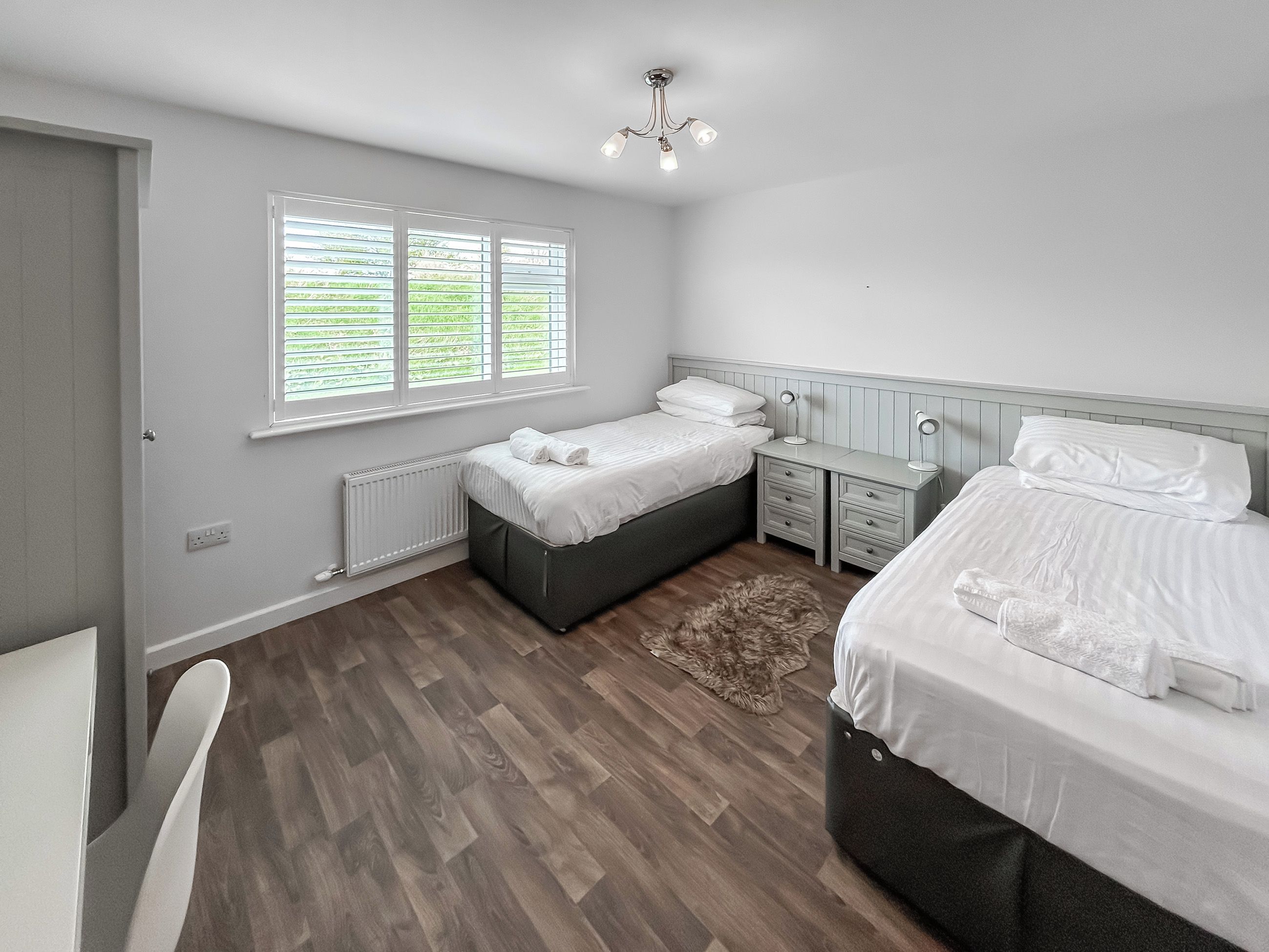 A bedroom with two beds, a bedside table, a chair, a radiator, and a window with blinds.