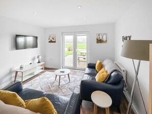 A modern living room with a sofa, a coffee table, a TV, and a sliding glass door leading to an outdoor area. - Primrose Lodge - UK51602 (Sunset Lakes)