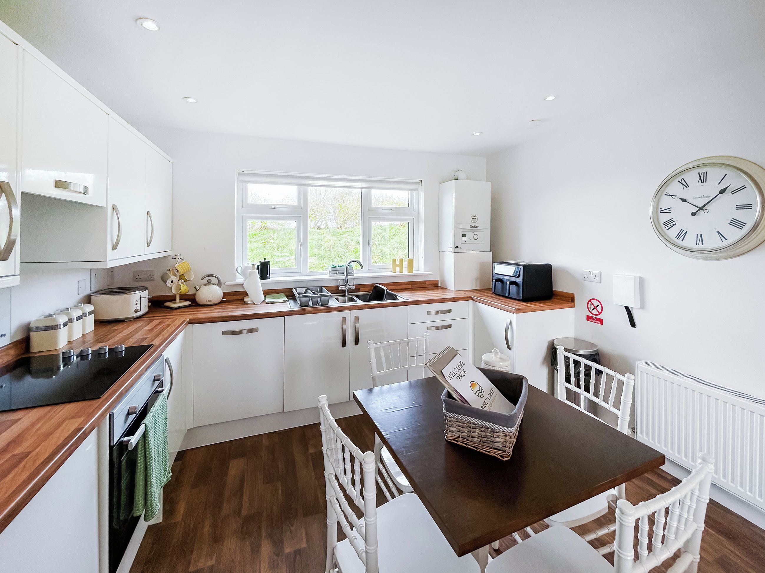 A modern kitchen with white cabinets, a wooden countertop, a dining table with chairs, a clock, and a window.