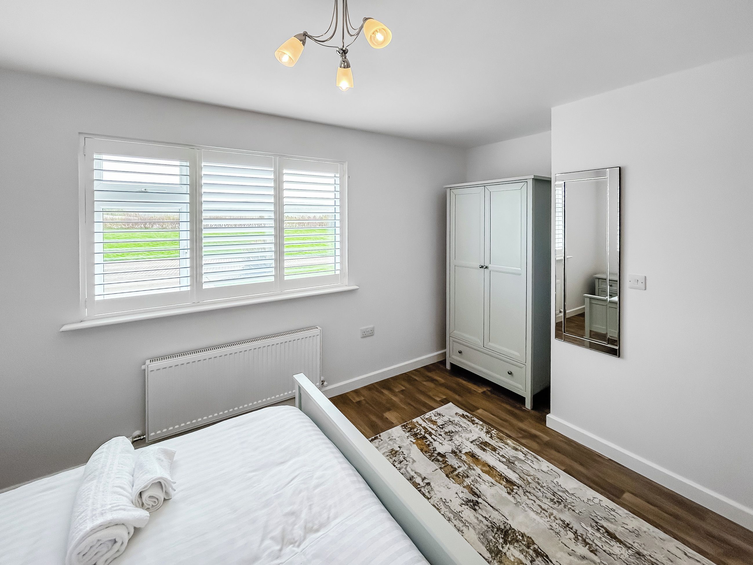 A bedroom with a bed, a radiator, a wardrobe, a mirror, and a window with blinds.