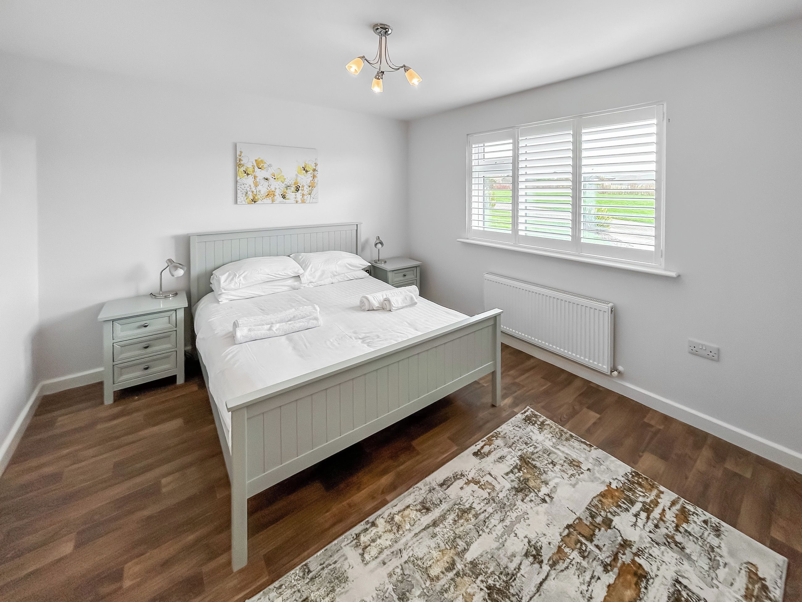 Une chambre à coucher comprenant un lit, des tables de chevet, un radiateur, une fenêtre avec des stores et un luminaire au plafond.