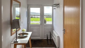 Un pasillo con una puerta blanca, un espejo, una mesa con un cuenco, una lámpara y vistas a un paisaje verde.