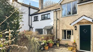 Une maison à deux étages, à l’extérieur blanc, avec une porte verte et une enseigne indiquant « Ash La Cottage ». Des plantes en pot sont disposées dans le jardin.