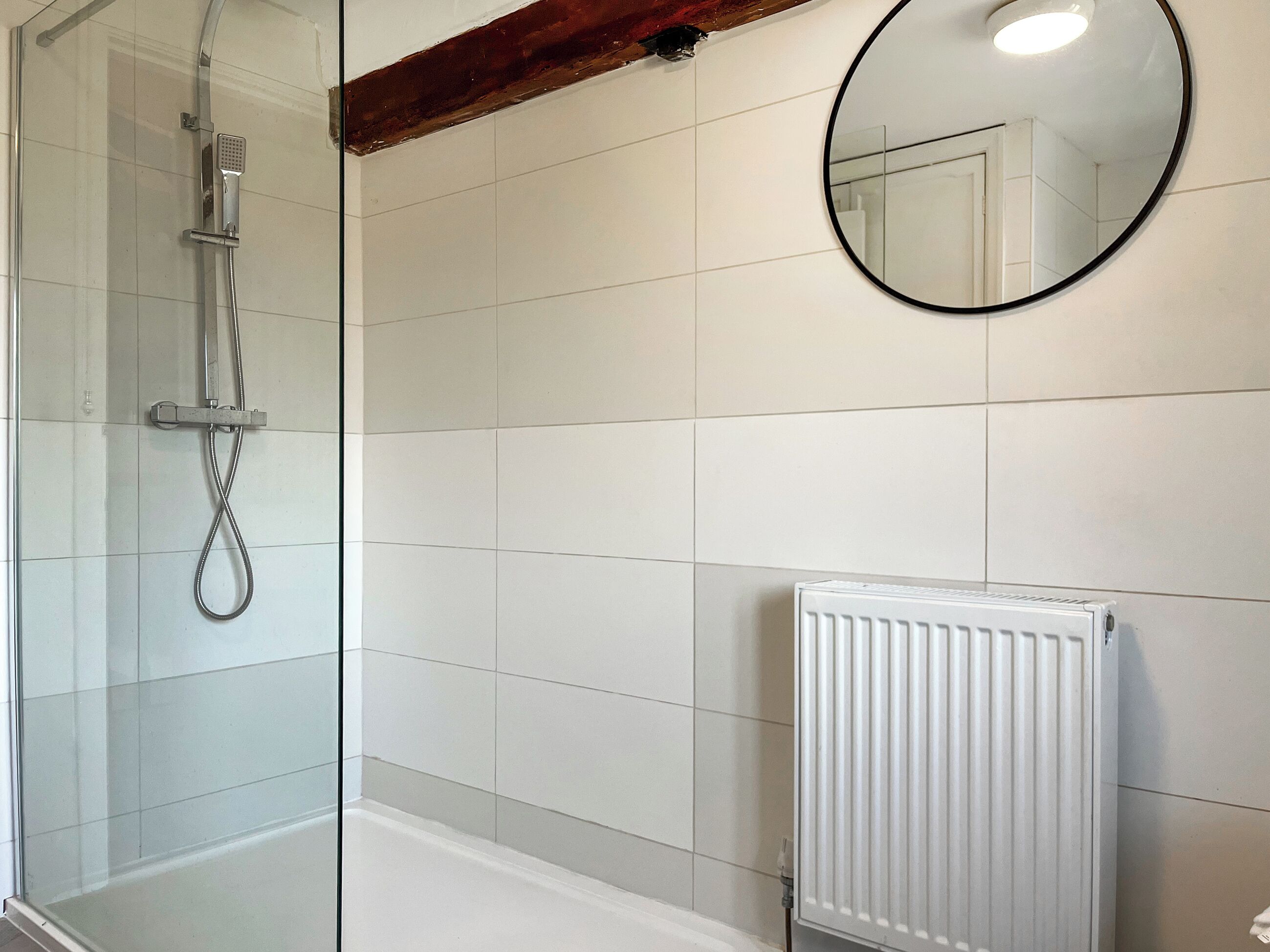 A bathroom with a glass shower enclosure, a white radiator, and a round mirror on the wall.