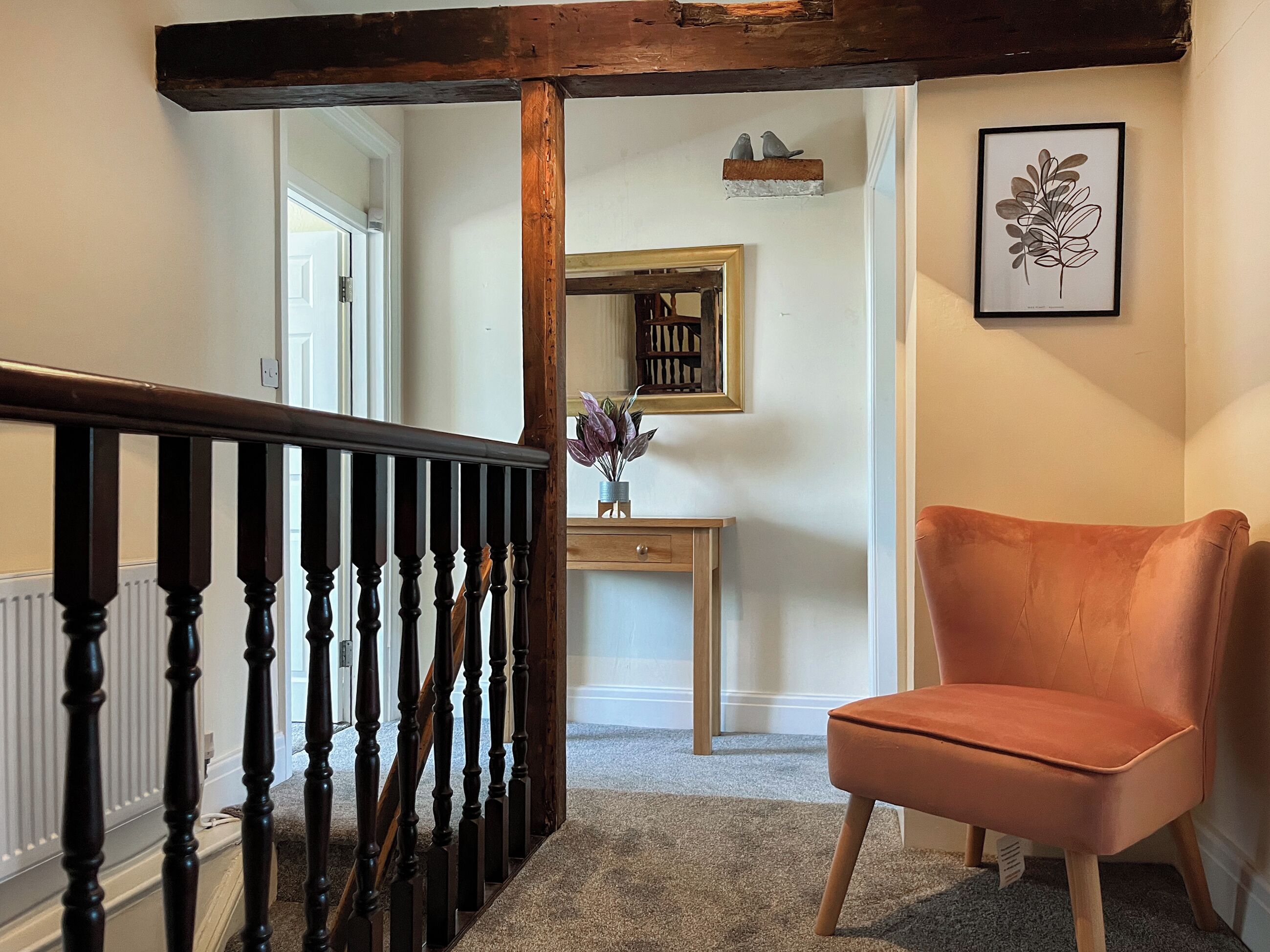 A staircase with a wooden handrail, a wooden desk with a mirror, a framed plant, and a cushioned chair.