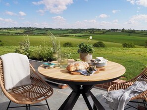 Een ronde houten tafel met stoelen, een boek, een glas, een broodje en een bloempot op een terras met uitzicht op groene velden.