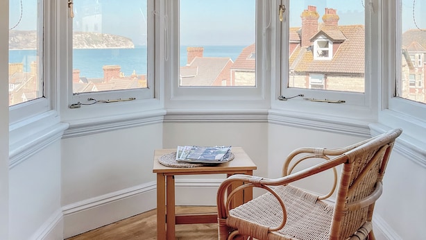Ett hörnrum med ett stort fönsterparti som vetter mot havet, ett träbord med tidningar och en fåtölj i rotting.