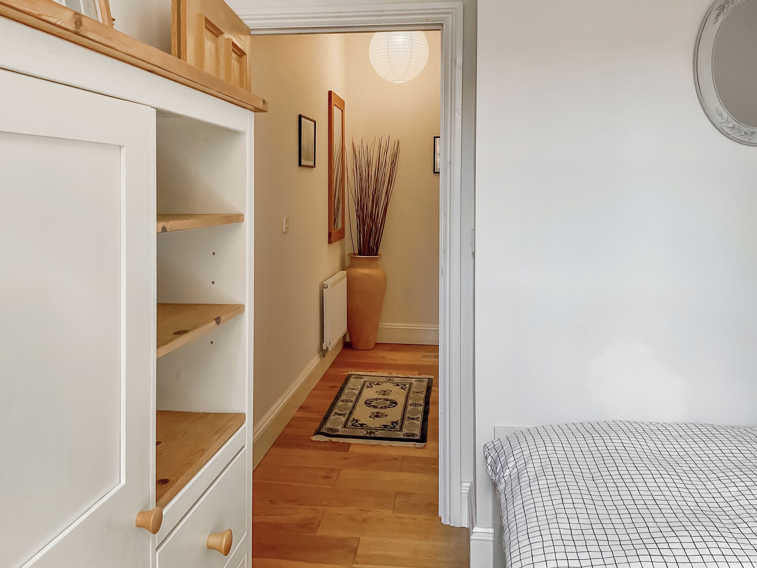 Un corridoio con un armadio bianco, un termosifone, un vaso con dei bastoncini, un tappeto decorato e un letto con copripiumino a quadretti.