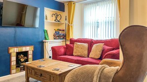 Ett vardagsrum med en röd soffa, ett soffbord i trä, en öppen spis, en tv som är monterad på väggen och en bokhylla.