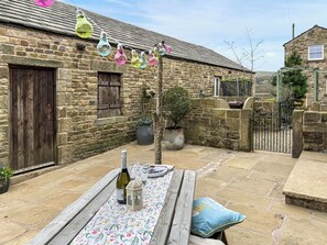 A stone building with a wooden door, a bench with a bottle and lantern, and a table with a floral tablecloth. - 3 bedroom accommodation in Chisworth (Chisworth)