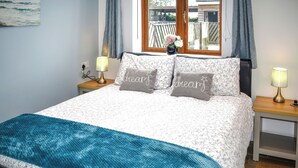 A neatly made bed with floral bedding, two bedside lamps, and a window with curtains. - 1 bedroom accommodation in Tytherleigh, near Axminster (Tytherleigh, near Axminster)