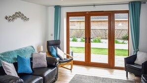 A living room with a black leather sofa, a wooden rocking chair, a grey rug, and a sliding glass door leading to a garden. - 1 bedroom accommodation in Tytherleigh, near Axminster (Tytherleigh, near Axminster)