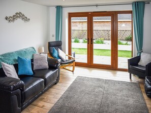Ein Wohnzimmer mit einem schwarzen Ledersofa, einem hölzernen Schaukelstuhl, einem grauen Teppich und einer Schiebetür, die in einen Garten führt.