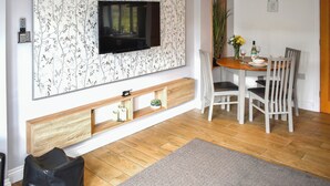 A living room with a flat-screen TV mounted on a wall with a floral wallpaper, a dining table with chairs, a small plant, and a wooden bench. - 1 bedroom accommodation in Tytherleigh, near Axminster (Tytherleigh, near Axminster)