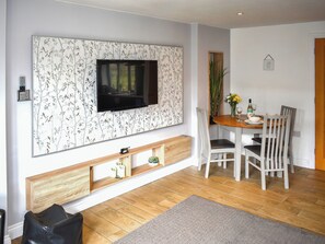 Ein Wohnzimmer mit Flachbildfernseher an einer Wand mit floralem Wandpapier, einem Esstisch mit Stühlen, einer kleinen Pflanze und einer hölzernen Bank.