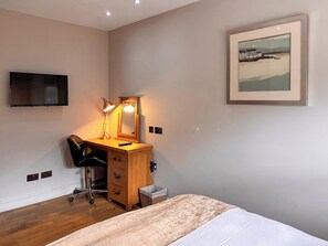A bedroom with a bed, a desk with a lamp, a chair, a TV, and a framed picture on the wall.