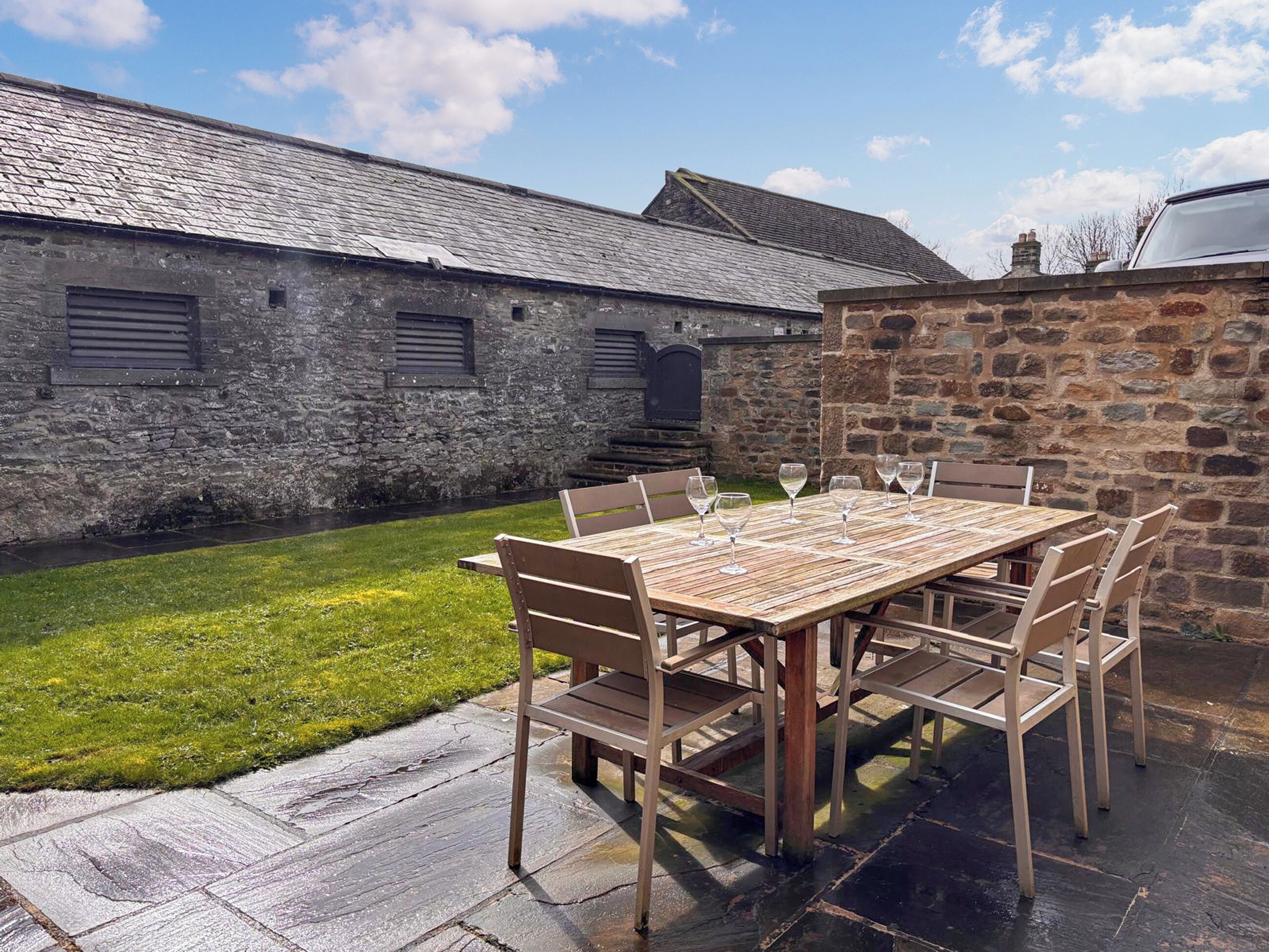A rustic outdoor dining area with a stone wall, a wooden table, and chairs.