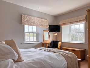 A bedroom with a bed, a desk, a chair, a television, and two windows with curtains.