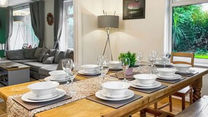 A dining area with a long wooden table set for six, a sofa in the background, and a view of greenery outside. - 5 bedroom accommodation in Pudsey, near Leeds (Pudsey, near Leeds)