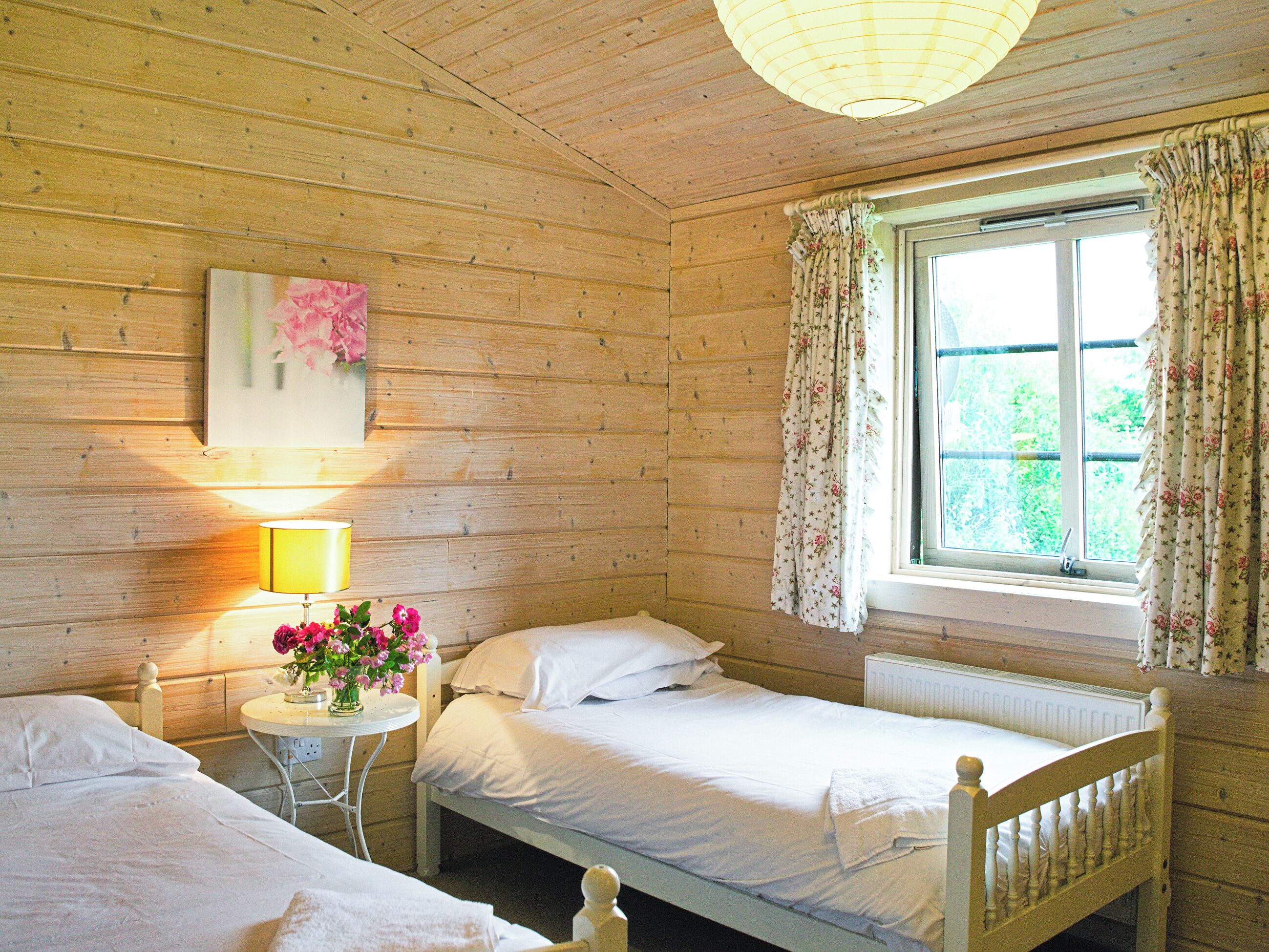 A room with wooden walls, two beds, a small table with a lamp, and a window with floral curtains.