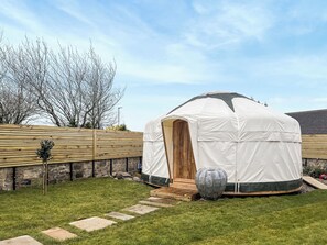 A white tent with a wooden door, set in a grassy yard with stone walls and a wooden deck.