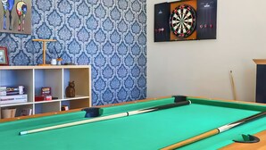A pool table with a dartboard and shelves with books and a cat figurine.