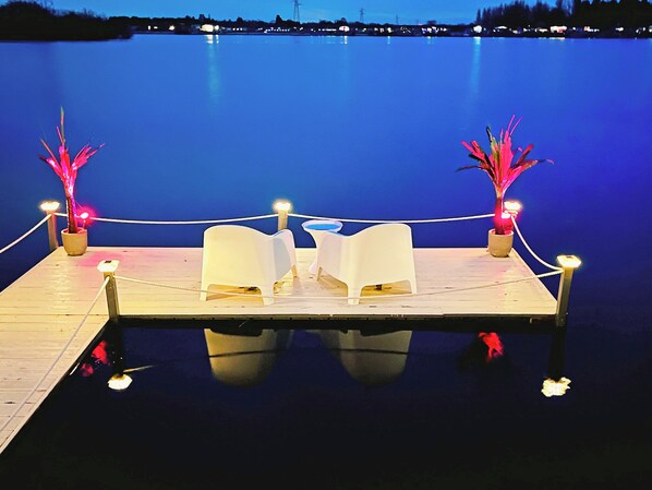 A dock with two white chairs and two potted plants with lights, overlooking a calm body of water.