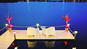 Un muelle con dos sillas blancas y dos plantas en macetas con luces, con vista a una masa de agua tranquila.