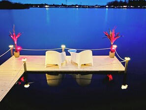 A dock with two white chairs and two potted plants with lights, overlooking a calm body of water.