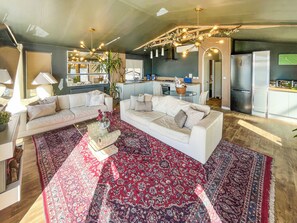 A modern living room with a large red patterned rug, two white sofas, a glass coffee table, and a kitchen area with a refrigerator and stove.