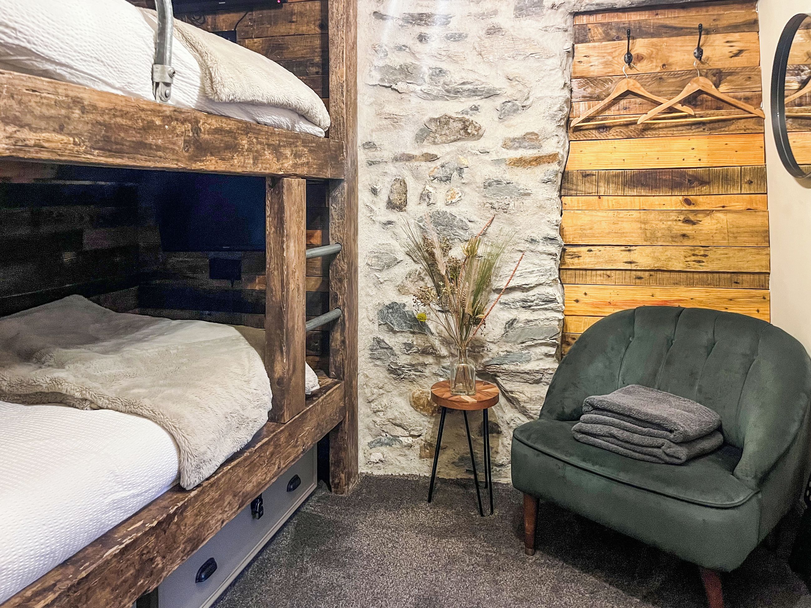 Een rustieke kamer met een stapelbed, een groene fauteuil, een klein houten tafeltje met een vaas en een stenen muur.