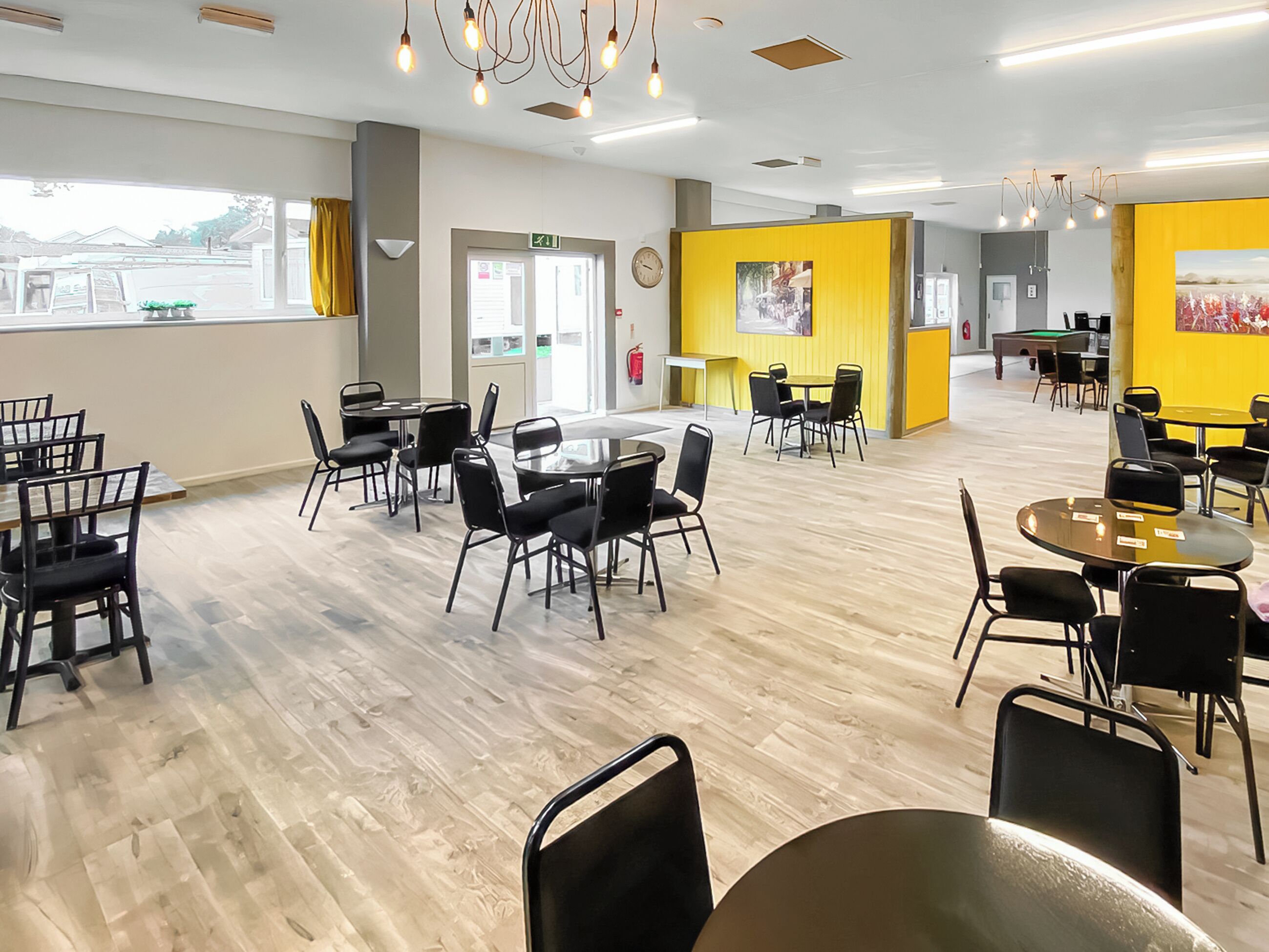 A spacious indoor area with multiple tables and chairs, a clock on the wall, and a yellow accent wall.