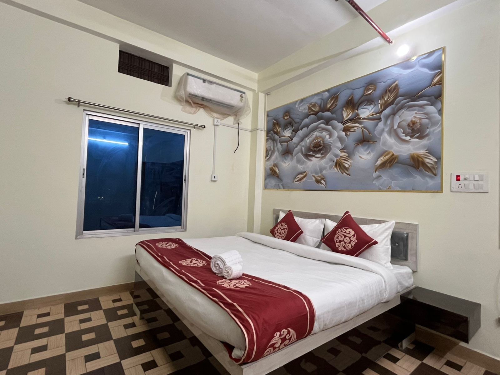 A hotel room with a bed, a window, an air conditioning unit, and a floral wall decoration.