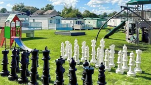 Un espacio con césped, un tablero de ajedrez, un parque infantil y casas móviles al fondo.