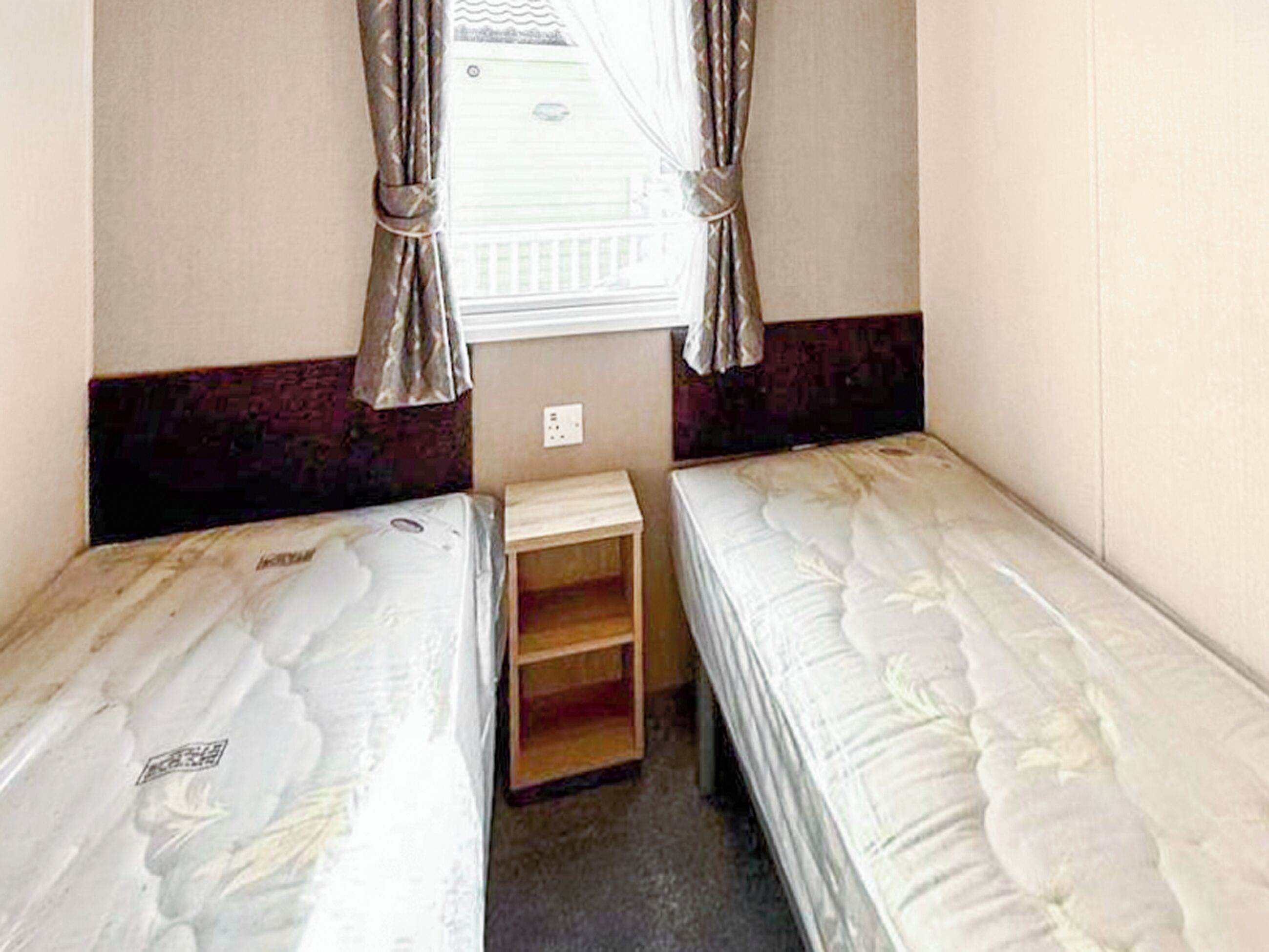 A small, compact room with two single beds, a small wooden shelf, and a window with curtains.