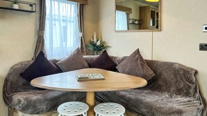 Ein gemütlicher Raum mit einem runden Holztisch, zwei weißen Hocker, einer Couch mit gemusterten Kissen, einem Spiegel und einem Fenster mit Vorhängen.