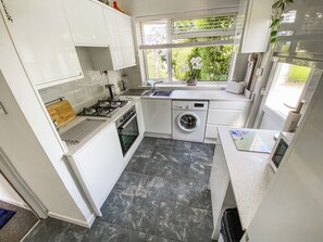A compact kitchen with white cabinets, a gas stove, a washing machine, and a window with a view of greenery. - 2 bedroom accommodation in Woolacombe (Woolacombe)
